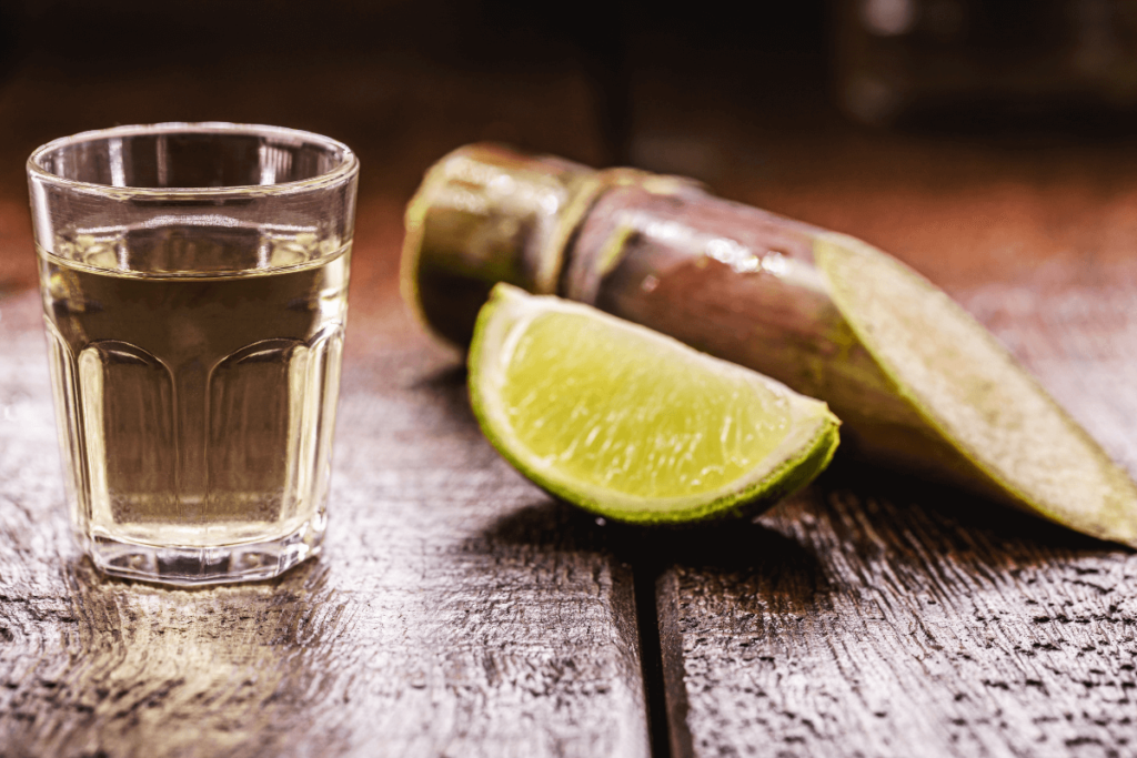 Um copo de cachaça em destaque, com um pedaço de cana-de-açúcar e uma rodela de limão ao fundo, sobre uma mesa de madeira rústica.