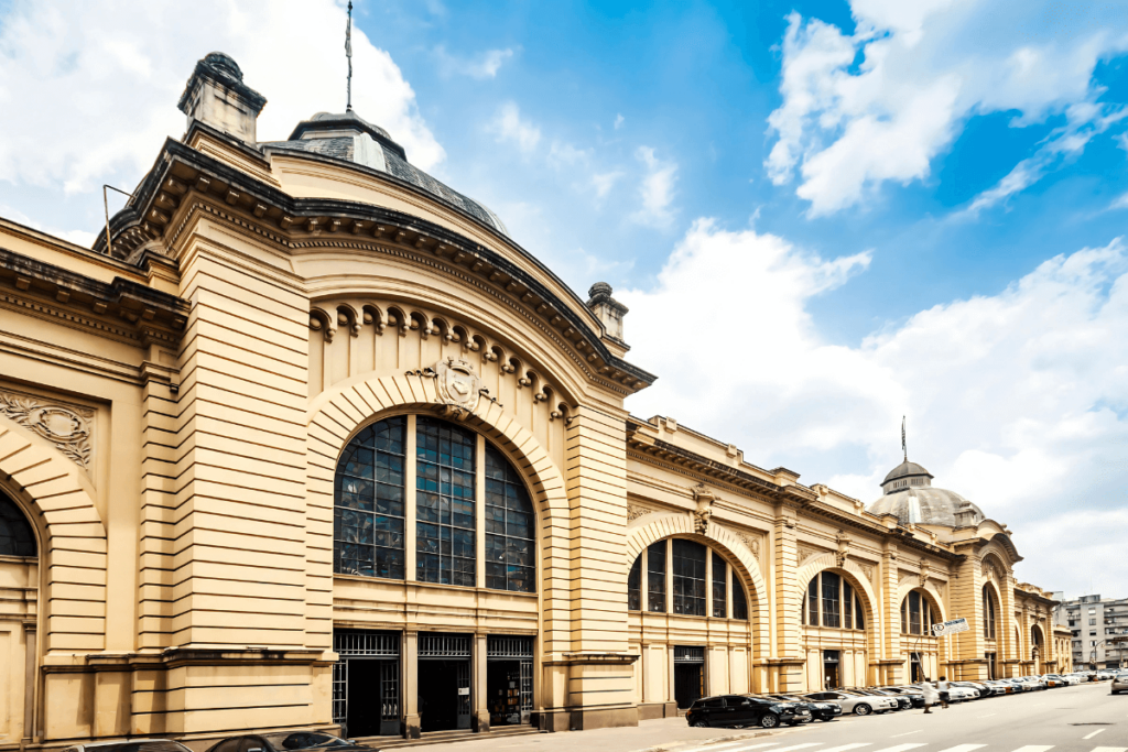 Fotografia exterior do Mercado Municipal de São Paulo, um marco da cozinha brasileira. Janelas em arco e arquitetura de fachada amarela. Marco para comida e ingredientes tipicos brasileiros.