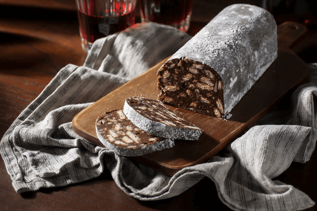 Palha italiana em formato de salame, polvilhada com açúcar, cortada em fatias e servida em tábua de madeira, destacando a textura e os ingredientes do doce brasileiro com história.