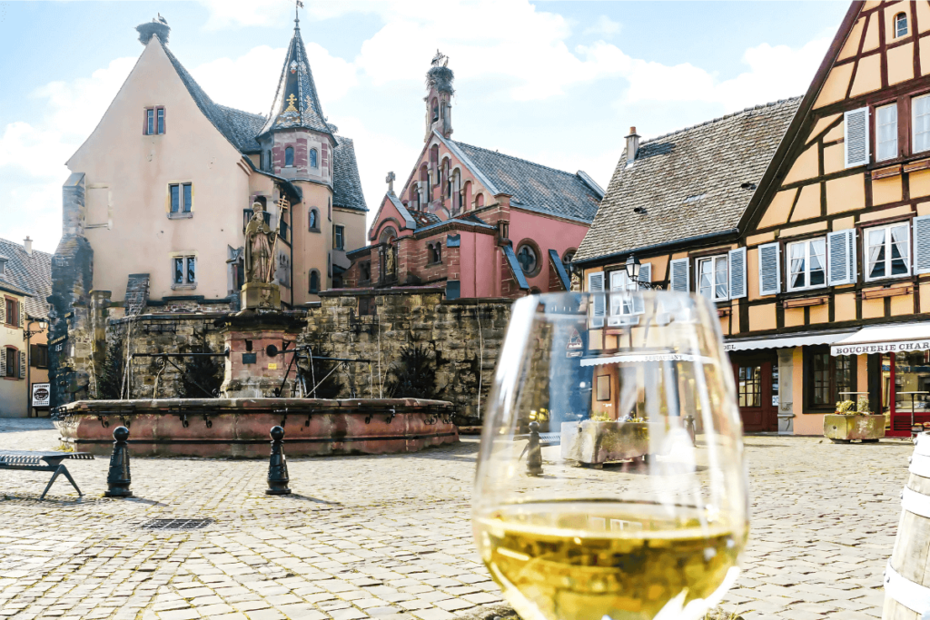 Taça de vinho branco em primeiro plano com vista para uma praça histórica com fonte em vilarejo da Alsácia.