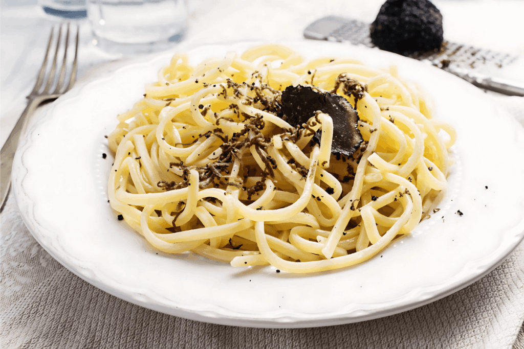 Prato de massa fresca Strangozzi coberto com raspas de trufa negra, um ingrediente requintado da Gastronomia do Centro da Itália, apresentado em um prato branco com um garfo e um ralador com uma trufa ao lado.