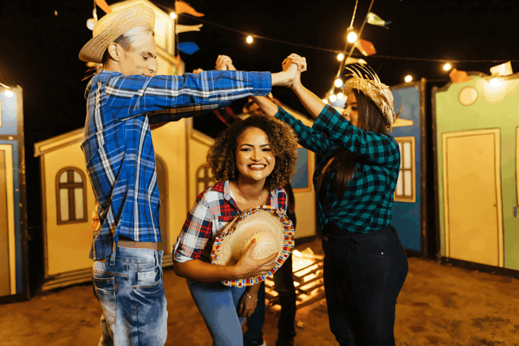 Três jovens sorrindo e dançando a tradicional quadrilha em uma festa junina. Eles usam chapéus de palha e camisas xadrez, em um arraial com casinhas típicas e luzes de festa, representando uma das danças.