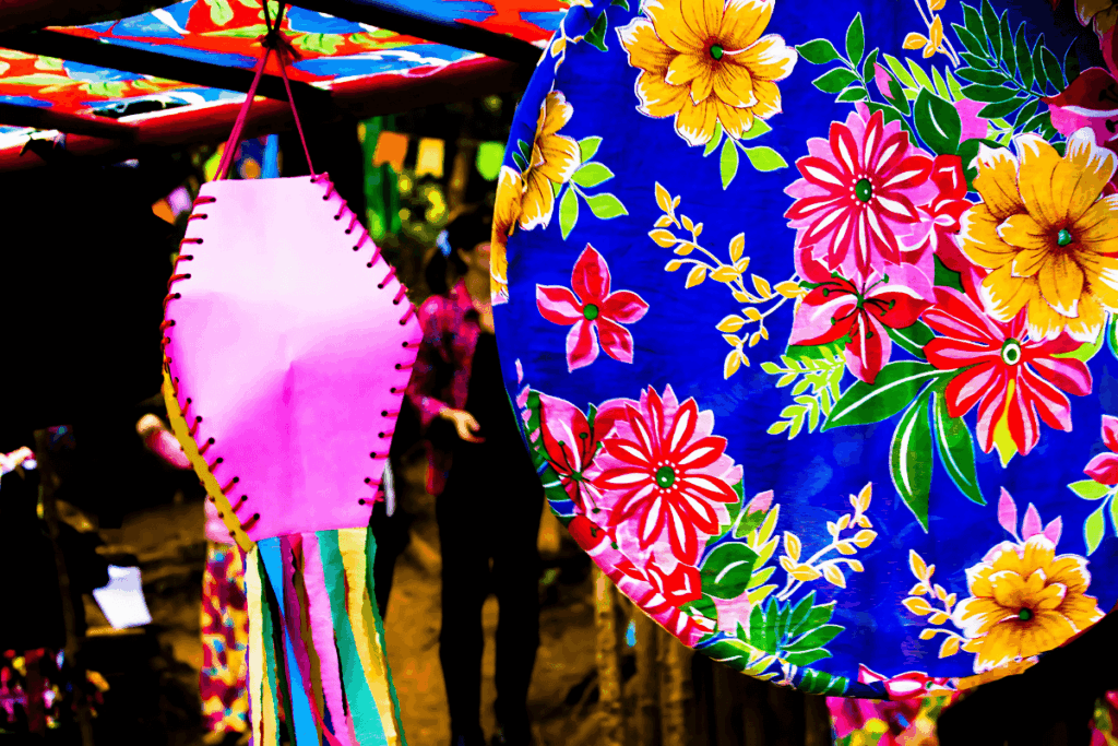 Close-up da decoração de uma festa junina tradicional, mostrando um balão de papel rosa pendurado ao lado de um enfeite azul com estampa floral vibrante. As cores fortes e o artesanato caracterizam o ambiente da festa.