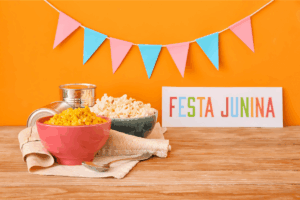 Composição minimalista com ícones de festa junina. Duas tigelas, uma com pipoca e outra com milho cozido, ao lado de uma placa colorida escrito 