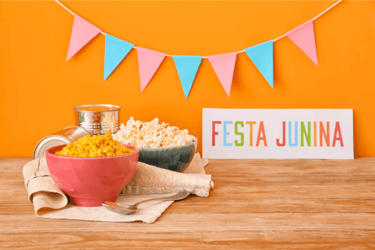 Composição minimalista com ícones de festa junina. Duas tigelas, uma com pipoca e outra com milho cozido, ao lado de uma placa colorida escrito 