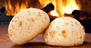 Dois pães de queijo dourados, ícones da culinária de Minas Gerais, sobre uma mesa de madeira com uma lareira acesa ao fundo.
